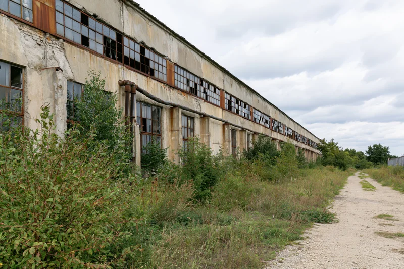 Long abandoned soviet-era industrial factory building with broken windows and overgrown vegetation along a dirt path