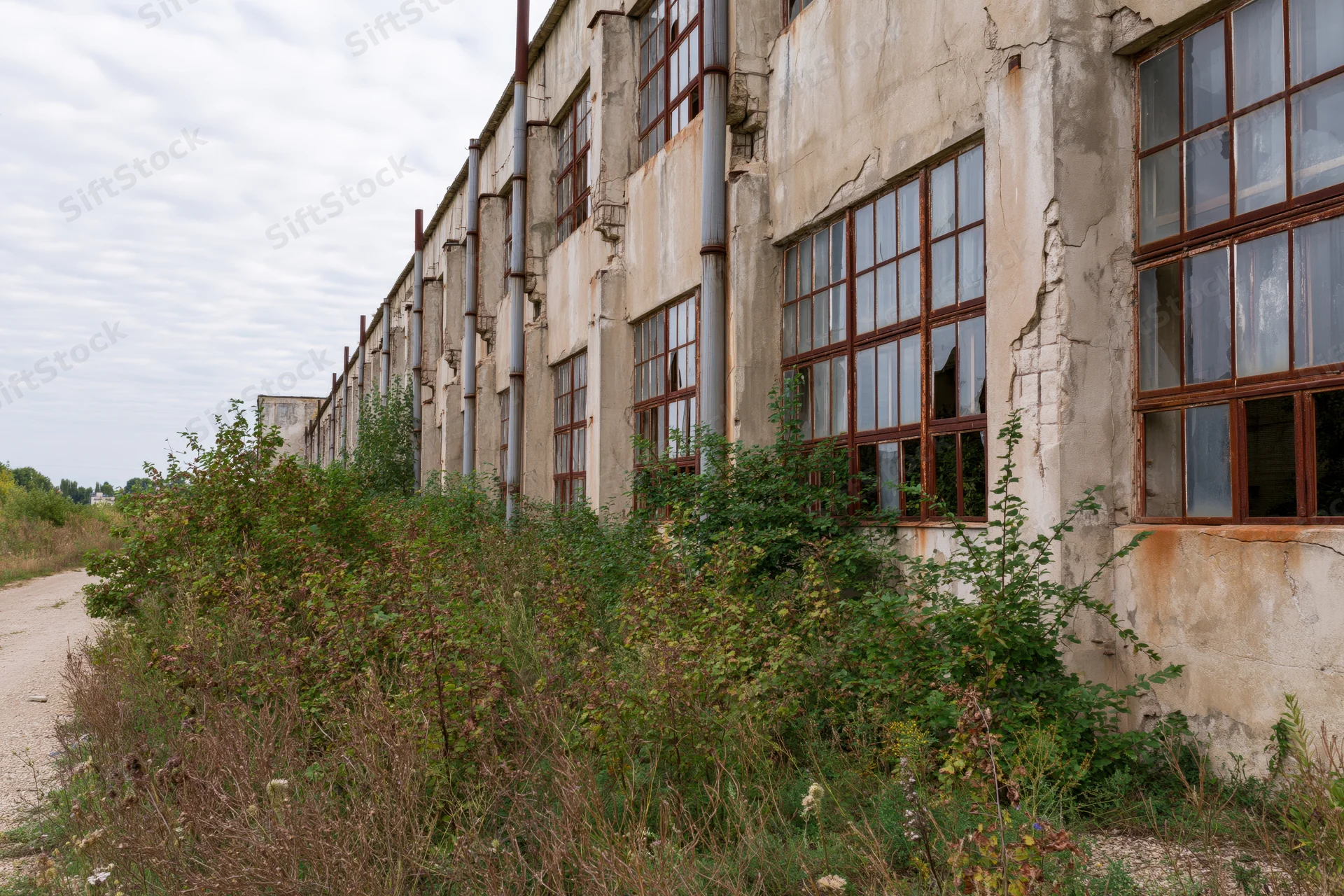Abandoned soviet-era industrial factory building with overgrown vegetation along its weathered concrete facade