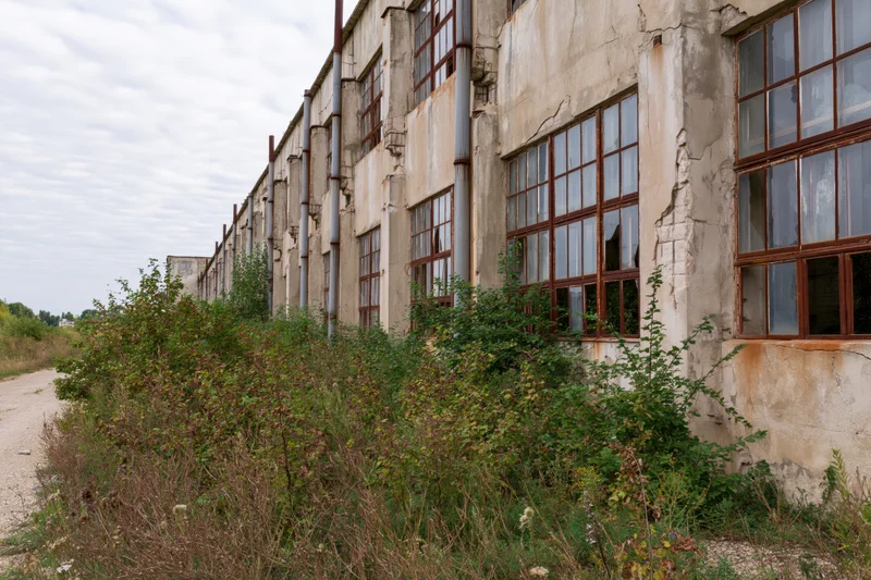 Abandoned soviet-era industrial factory building with overgrown vegetation along its weathered concrete facade
