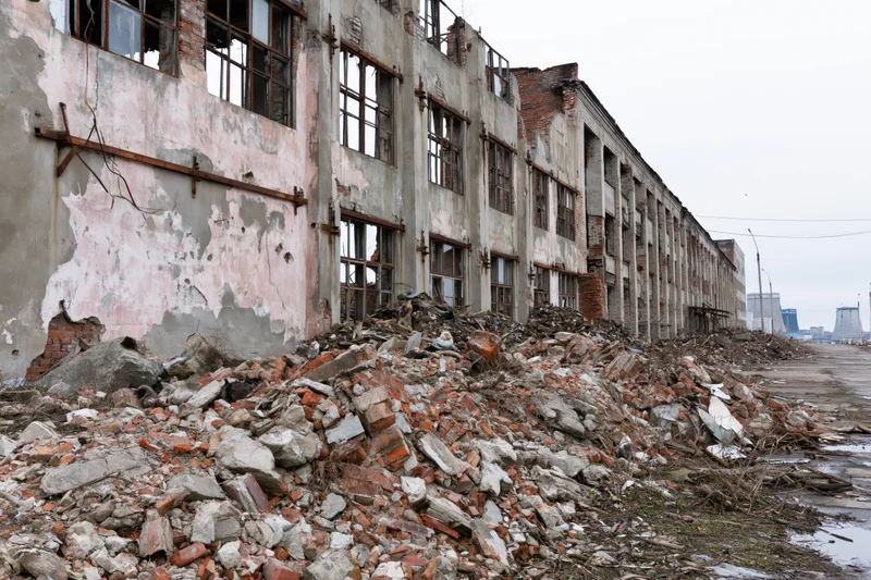 Abandoned soviet-era industrial factory building with crumbling walls and rubble pile along the side of a paved road