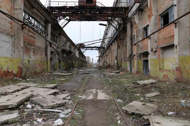 Abandoned soviet-era industrial factory complex with rusted gantry cranes and overgrown rail tracks between decaying brick buildings