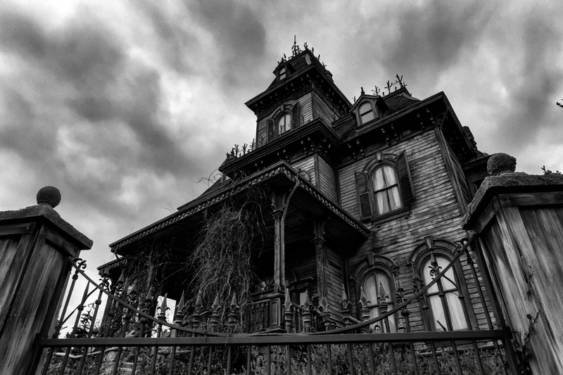 Abandoned victorian mansion with weathered wood and overgrown vines under stormy sky, viewed from low angle through iron gate