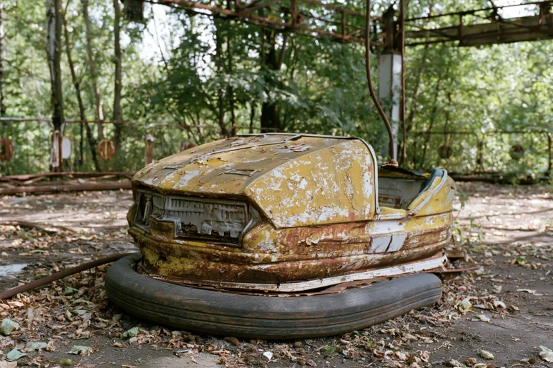 Decayed yellow bumper car resting on dirt floor surrounded by overgrown trees and foliage in abandoned amusement park