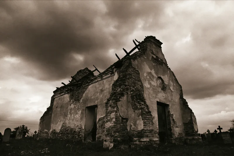 Crumbling stone chapel with collapsed roof under overcast sky in cemetery