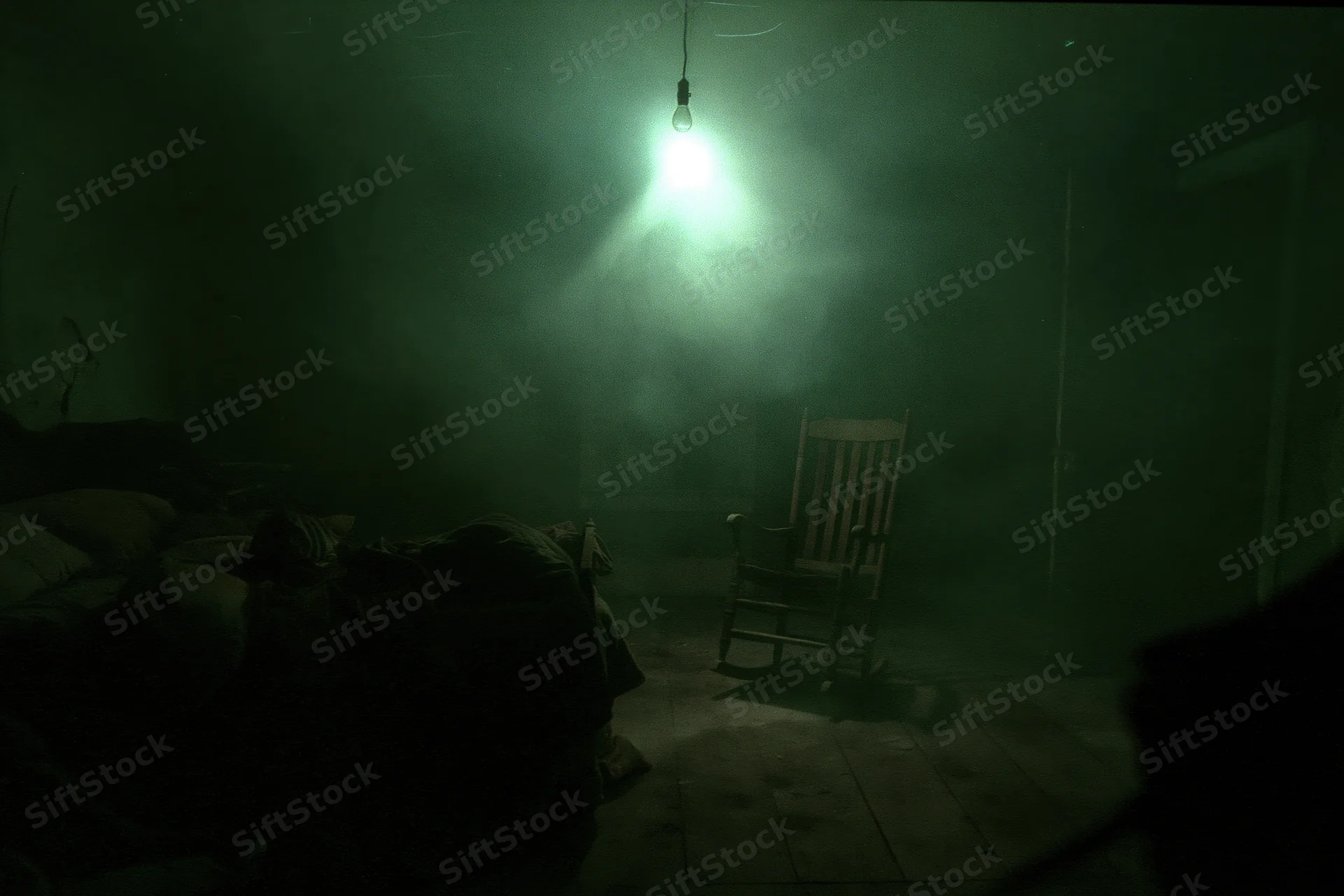 Dim green light from hanging bulb illuminates unmade bed and wooden rocking chair in abandoned dark bedroom