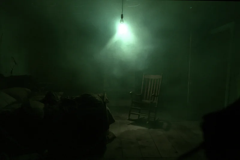 Dim green light from hanging bulb illuminates unmade bed and wooden rocking chair in abandoned dark bedroom