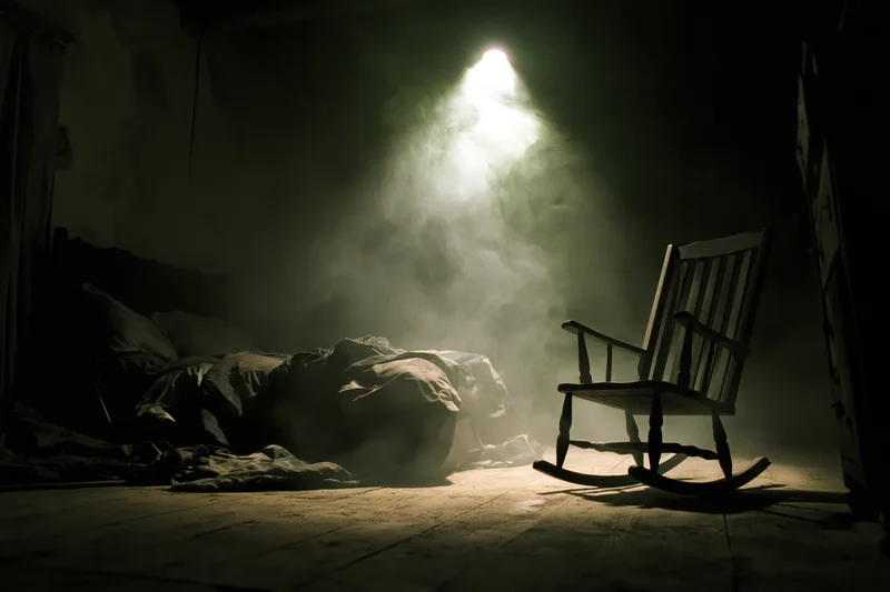 Dimly lit abandoned bedroom with unmade bed and wooden rocking chair in dusty haze