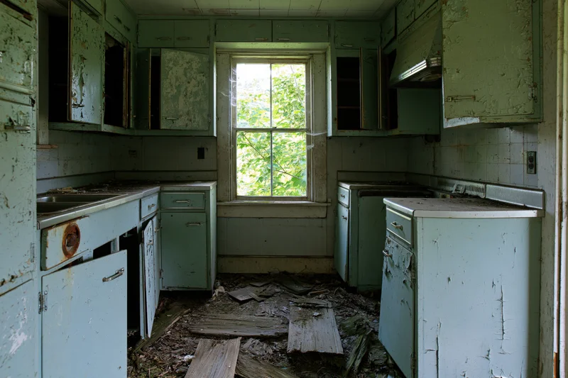 Abandoned farmhouse kitchen interior with peeling green cabinets and debris on the floor