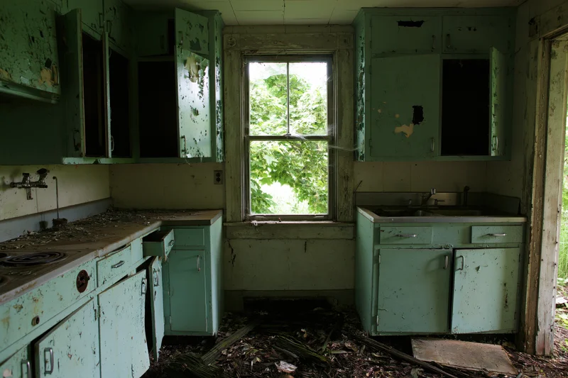 Abandoned farmhouse kitchen interior with peeling green cabinets and debris on the floor