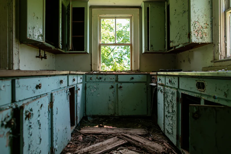 Abandoned farmhouse kitchen interior with peeling green cabinets and debris on the floor, natural light through window
