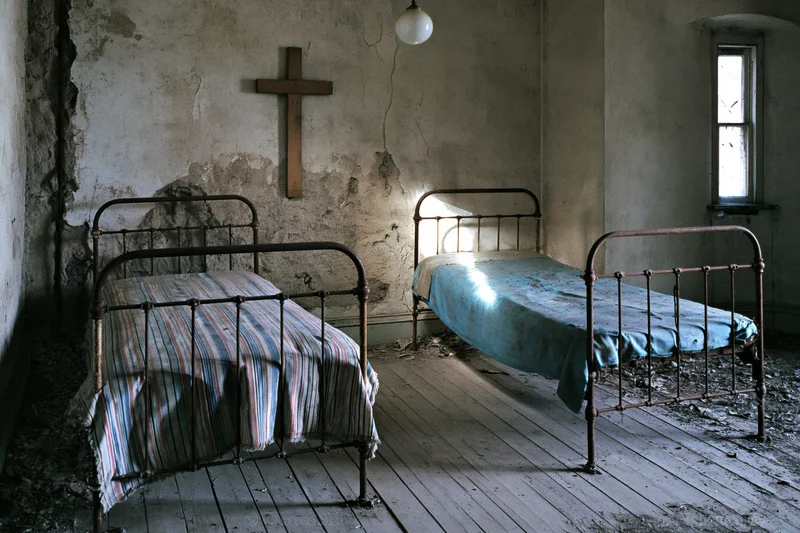 Two rusted iron beds in a derelict hospital room with a wooden cross on the wall and sunlight through a window