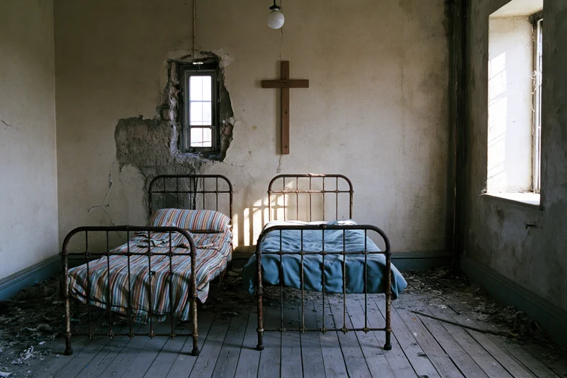 Two rusted iron beds in a decaying hospital room with a wooden cross on the wall