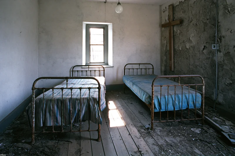 Two rusted iron beds in a dilapidated abandoned hospital room with peeling walls and a wooden cross