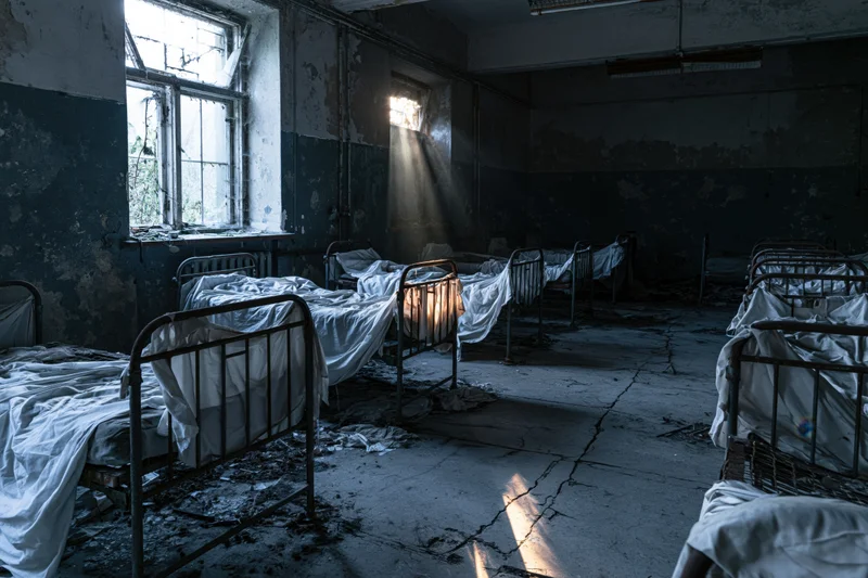 Rows of metal beds in an abandoned hospital ward with sunlight streaming through broken windows