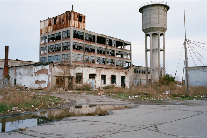 Abandoned industrial factory complex with crumbling multi-story building and water tower, overgrown with weeds and cracked pavement, puddle reflecting the structure