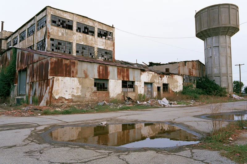 Abandoned industrial factory complex with crumbling buildings and a water tower, surrounded by overgrown vegetation and cracked pavement with puddles reflecting the structures