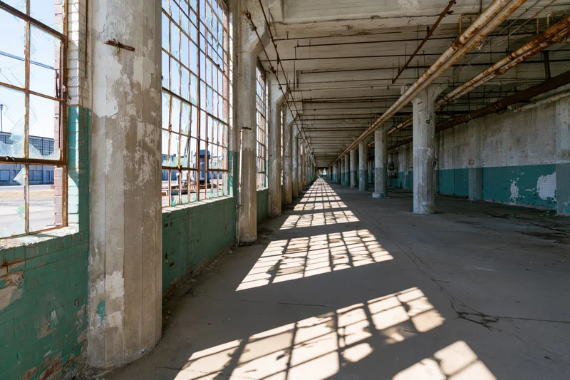 Long empty corridor in abandoned factory with sunlight streaming through large windows casting grid shadows on concrete floor