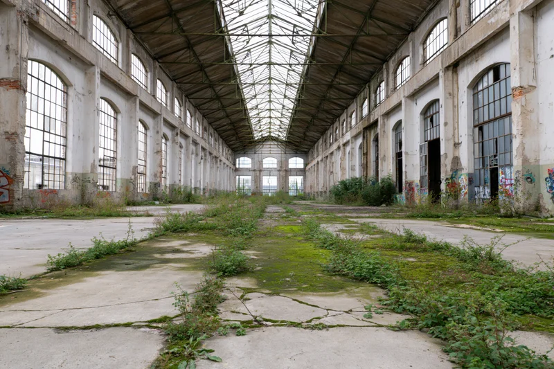 Long symmetrical view of abandoned industrial factory hall interior with overgrown vegetation and arched windows