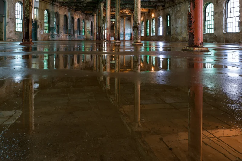 Abandoned industrial warehouse interior with wet concrete floor reflecting pillars and arched windows