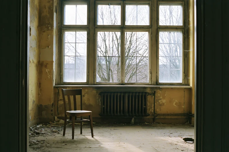 Single wooden chair in an abandoned room with peeling walls and a large window overlooking bare trees
