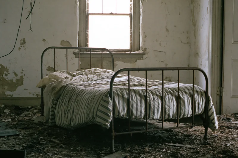 Abandoned iron bed frame with sagging striped mattress in a derelict room filled with debris