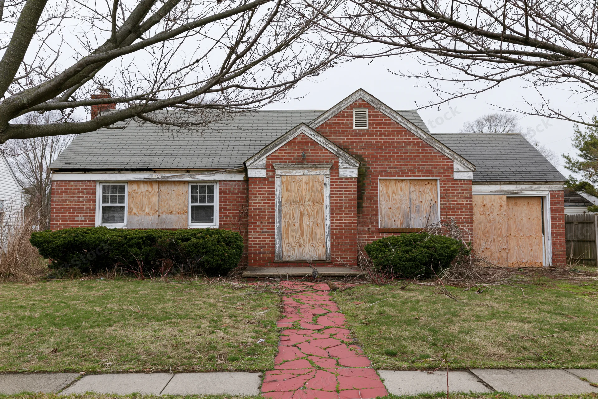 Abandoned brick cape cod house with plywood-covered windows and door in suburban yard