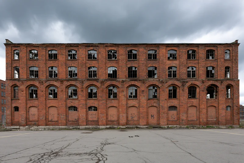 Abandoned red brick industrial factory building with broken windows and cracked pavement under overcast sky