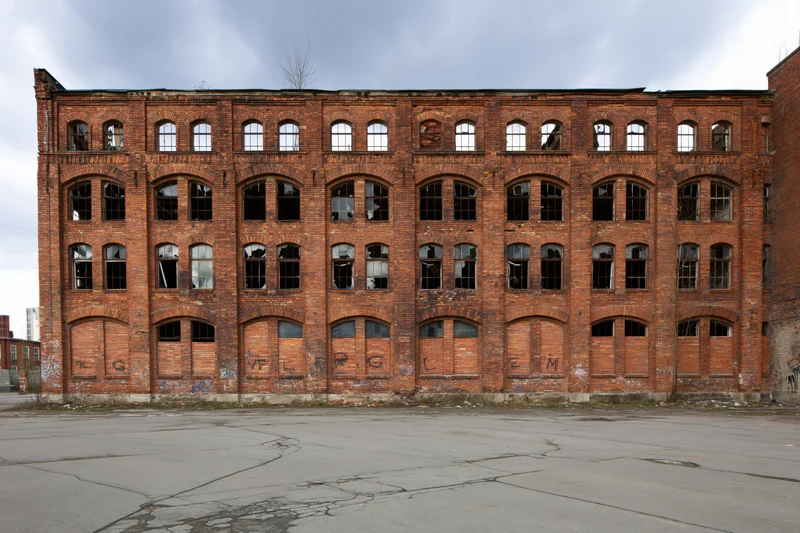 Abandoned red brick industrial factory building with broken windows and graffiti on facade under overcast sky