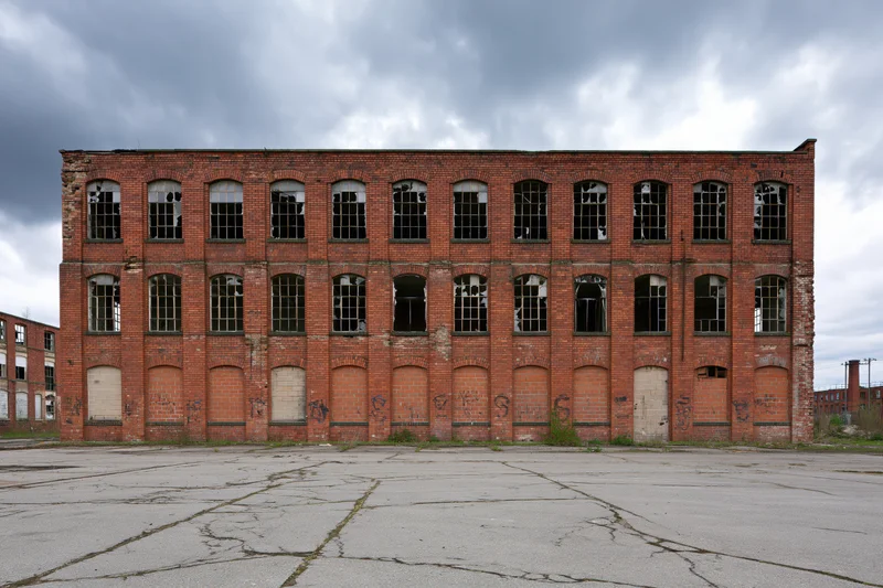 Abandoned red brick industrial factory building with broken windows and cracked pavement under overcast sky