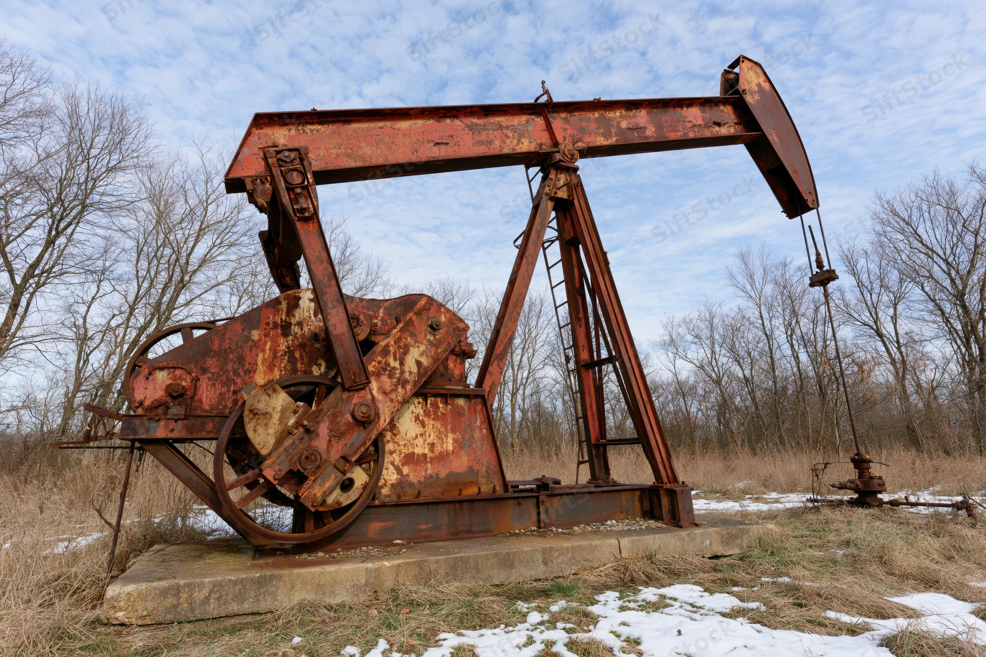 Abandoned rusty oil pump jack sitting idle on concrete foundation in snowy field