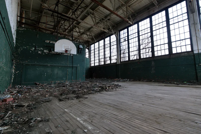Abandoned school gymnasium interior with decaying basketball hoop and debris on wooden floor