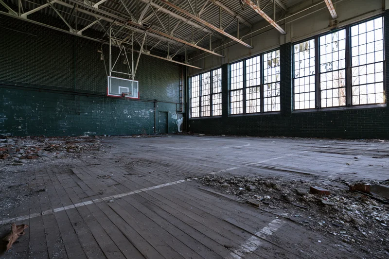 Abandoned school gymnasium interior with decaying wooden floor and broken windows
