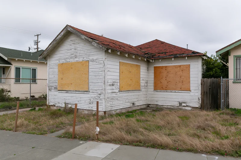 Abandoned single-story clapboard house with boarded-up windows and overgrown yard on a residential street