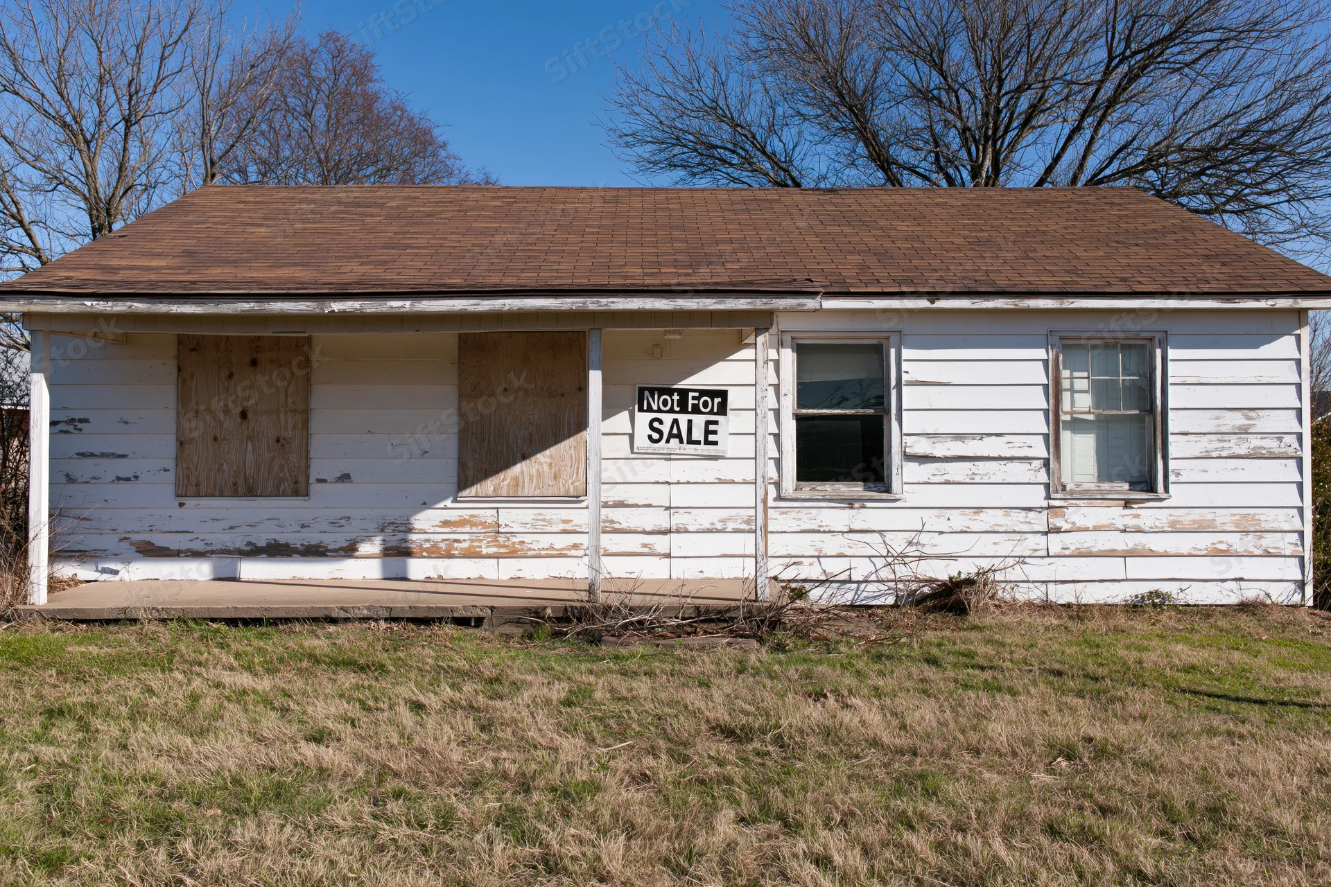 Abandoned single-story ranch house with boarded windows and not for sale sign on peeling white siding