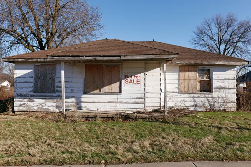 Abandoned single-story ranch house with boarded windows and not for sale sign on front porch