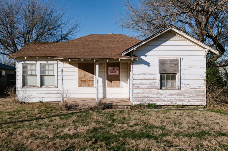 Abandoned single-story ranch house with boarded windows and not for sale sign on front porch