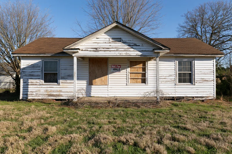 Abandoned single-story ranch house with boarded windows and not for sale sign on front porch in rural yard