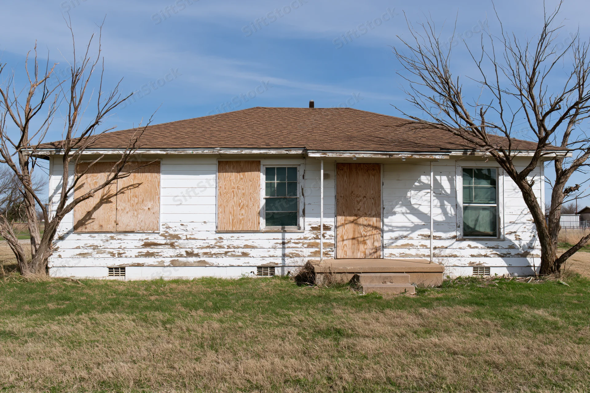 Abandoned single-story ranch house with plywood-boarded windows and peeling white siding in a rural yard