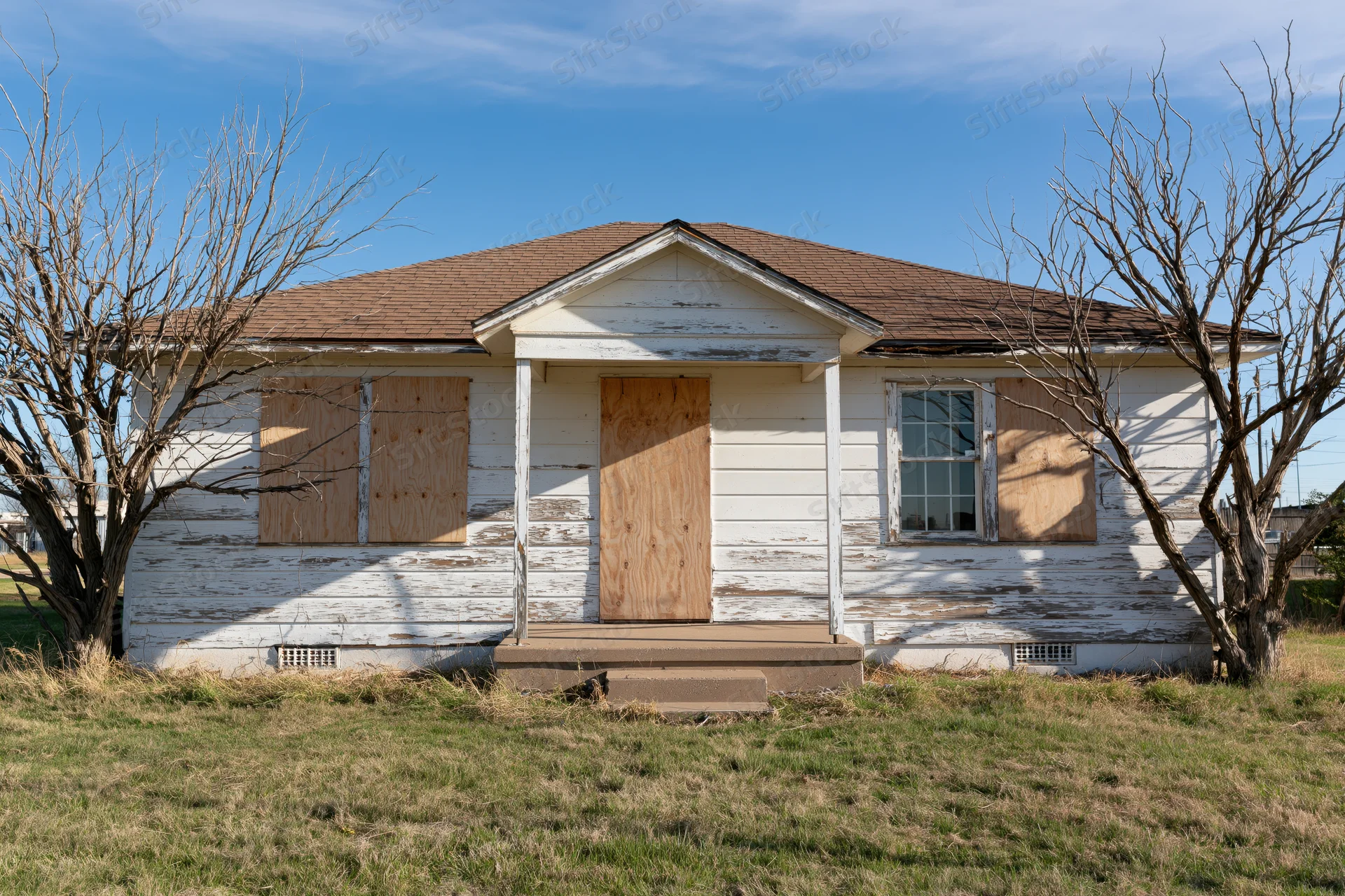 Abandoned single-story ranch house with plywood-boarded windows and door under a blue sky