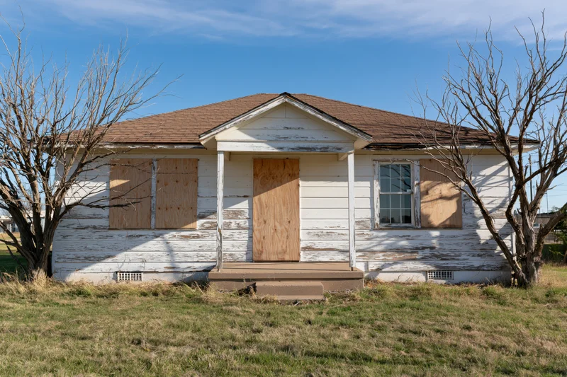 Abandoned single-story ranch house with plywood-boarded windows and door under a blue sky