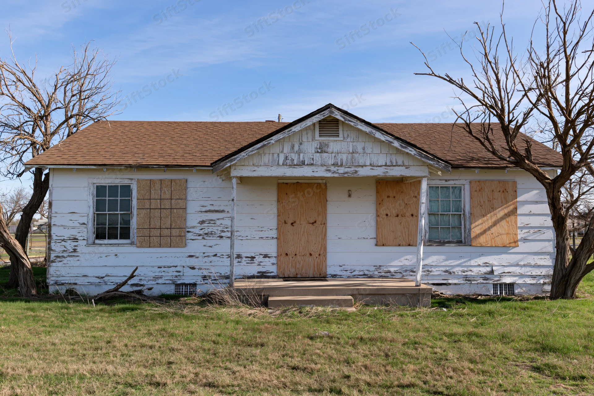 Abandoned single-story ranch house with plywood-boarded windows and weathered white siding in a rural yard