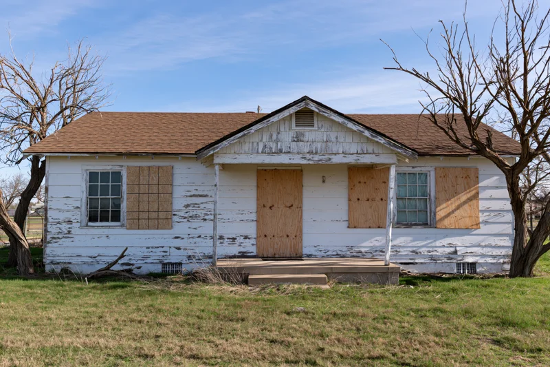 Abandoned single-story ranch house with plywood-boarded windows and weathered white siding in a rural yard
