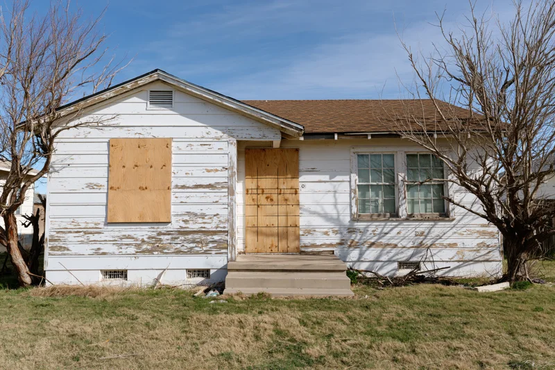 Abandoned single-story ranch house with plywood-boarded windows and door under a blue sky