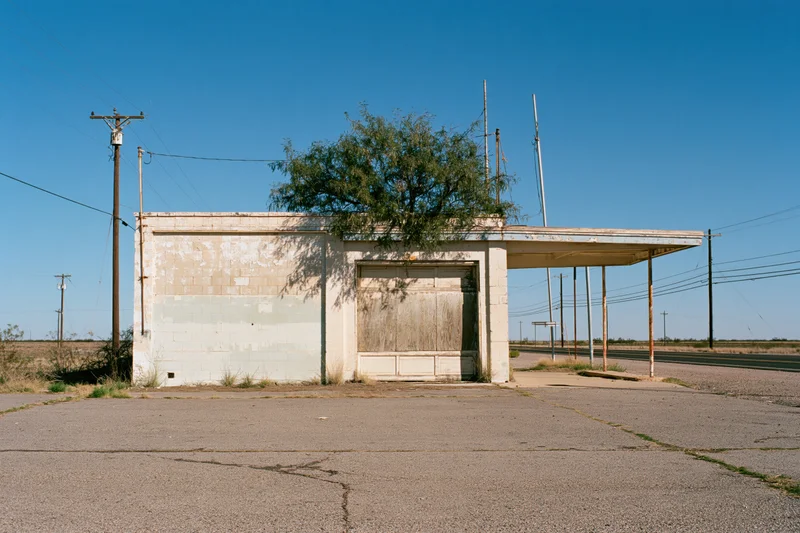 Abandoned single-story roadside service station with boarded windows and overgrown tree under clear blue sky
