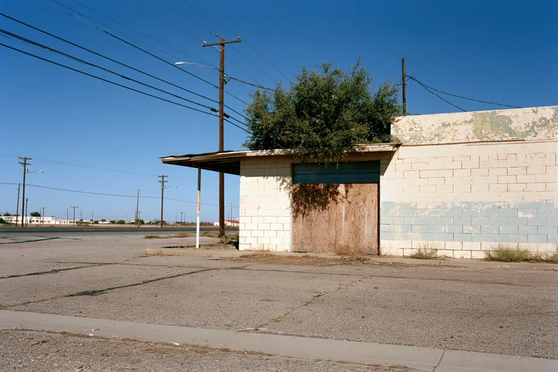 Abandoned single-story roadside service station with boarded door and overgrown vegetation under clear blue sky