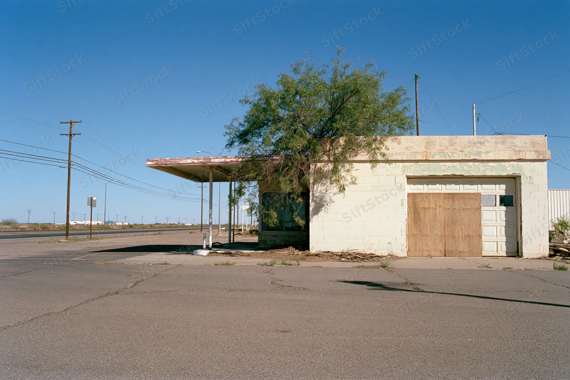 Abandoned single-story roadside service station with boarded garage door and overhang under clear blue sky