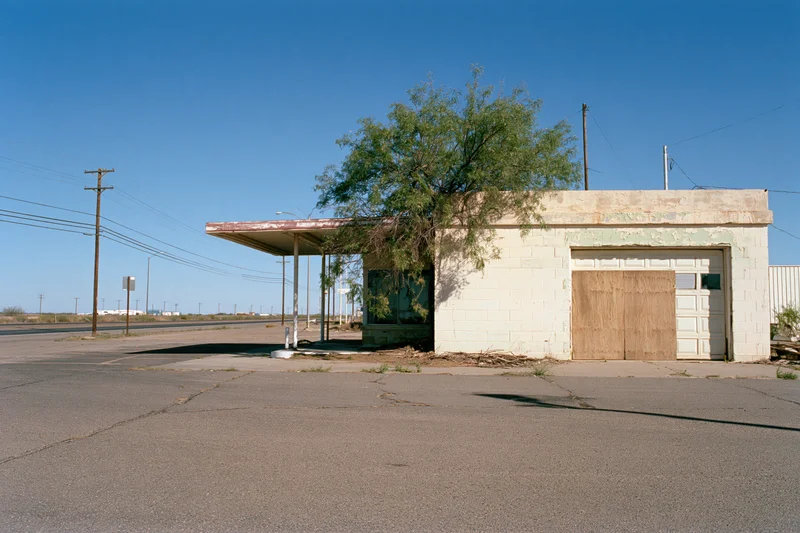 Abandoned single-story roadside service station with boarded garage door and overhang under clear blue sky