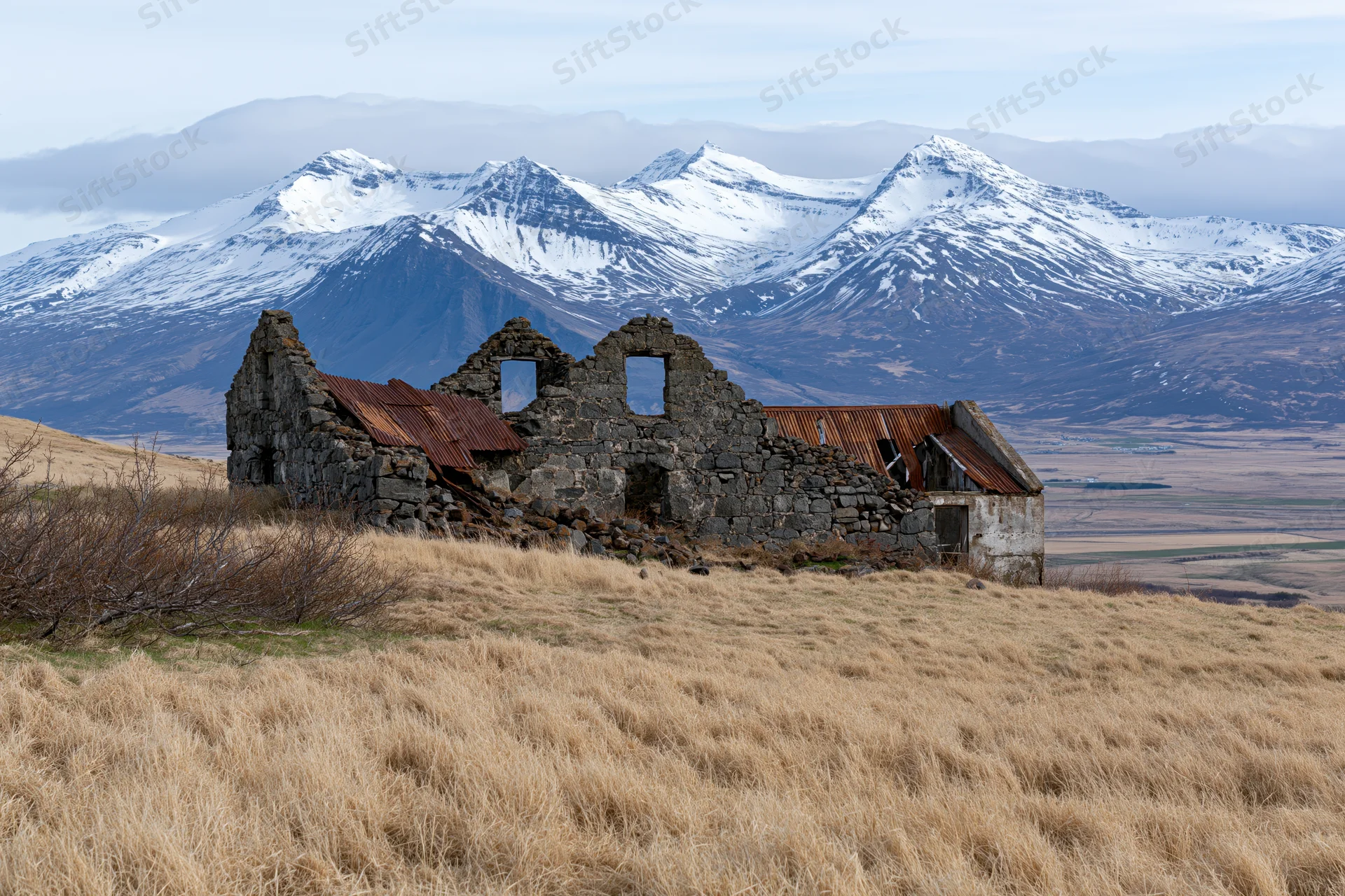 Abandoned stone farmhouse ruins on a grassy hill with snow-capped mountains in the background