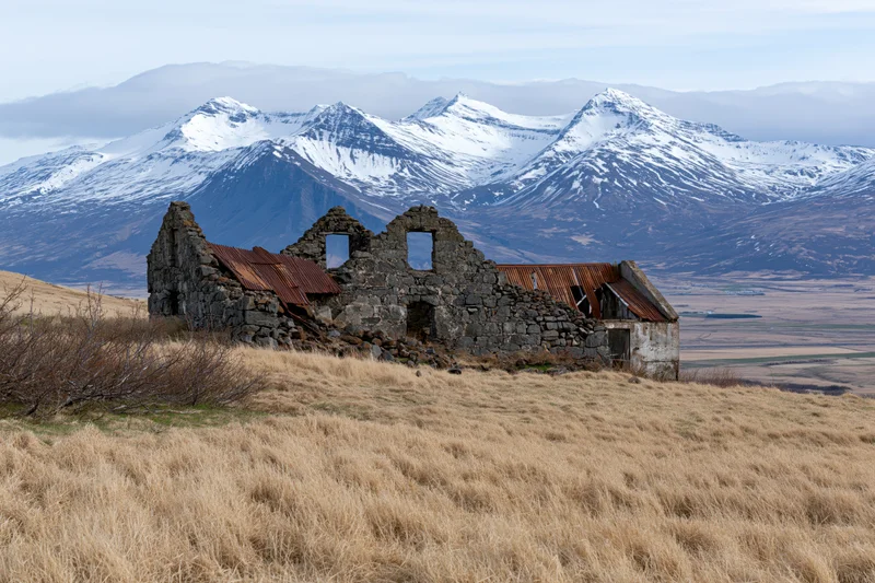 Abandoned stone farmhouse ruins on a grassy hill with snow-capped mountains in the background