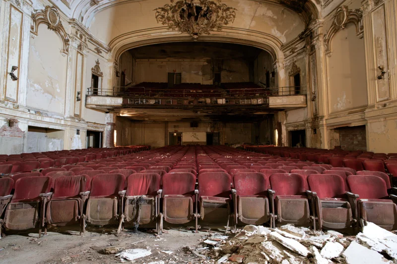 Rows of red seats in a decaying theater interior with broken plaster and peeling walls